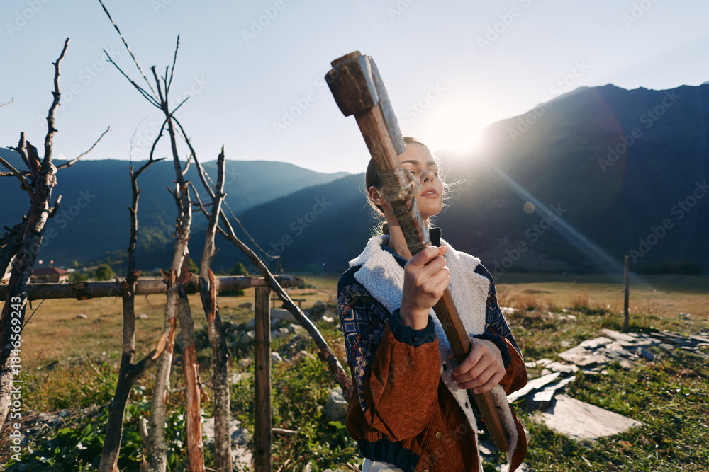 Fototapeta premium woman with axe in countryside near wooden fence and mountains with sun flare, outdoor rural scene of rustic woodcutting and manual work, portrait of determined person preparing firewood