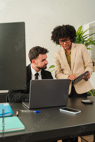Two professional colleagues actively collaborating while using a laptop in an office, showcasing teamwork and modern business practices. One individual is sharing insights on a tablet.