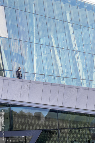 View of a lone figure stands on a stark white balcony against the backdrop of a modern glass building reflecting the sky, Oslo, Norway.