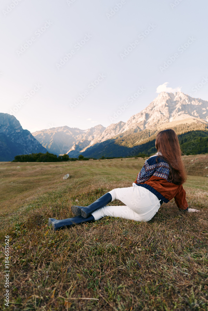 Fototapeta premium Woman in a meadow relaxing and watching mountains and dramatic landscape, nature escape with sweater and boots, outdoor travel scene in a peaceful alpine valley panorama.
