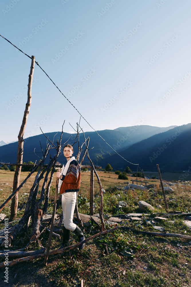 Fototapeta premium Woman portrait by rustic fence in a rural field with mountains in the background, natural landscape and sunlight. Outdoor nature scene with casual clothing and contemplative mood.