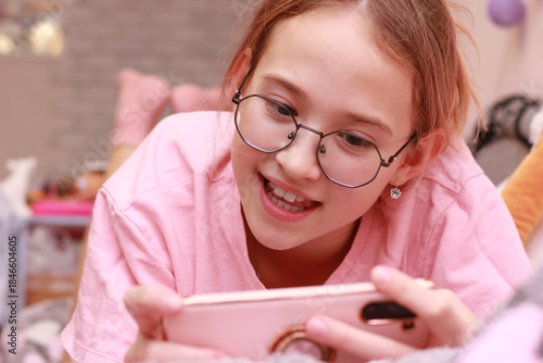 Close-up of a teenage girl, smiling, lying on a bed and playing a mobile game with a smartphone. Leisure in a cozy pink bedroom