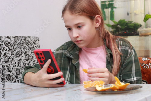 A teenage girl is sitting at the kitchen table and eating a tangerine. He's watching videos on his phone.
