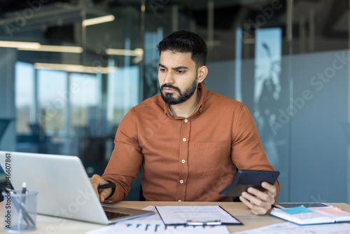 Young businessman or accountant checking financial data, using a calculator and laptop while working at an office desk, focused on managing business finances and economy strategy