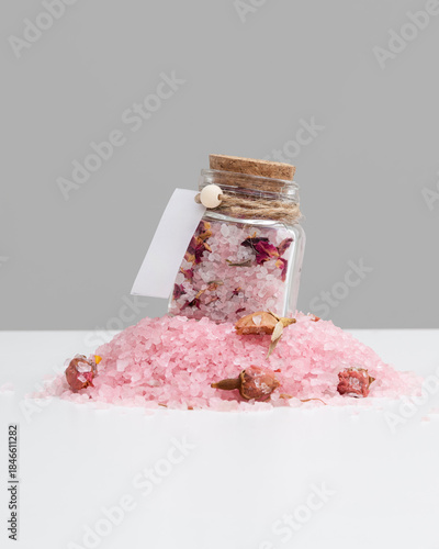 A small glass jar filled with pink sea salt and dried rose petals, perched on a mound of the same salt. The jar is adorned with a cork stopper and a blank white tag. Bonbonniere. mockup