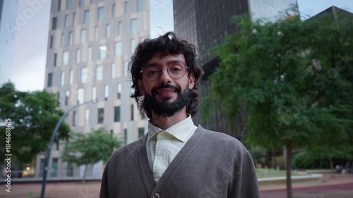 Smiling millennial professional with glasses stands in a modern business district, making eye contact with the camera. Urban office towers behind him suggest confidence, leadership and career ambition