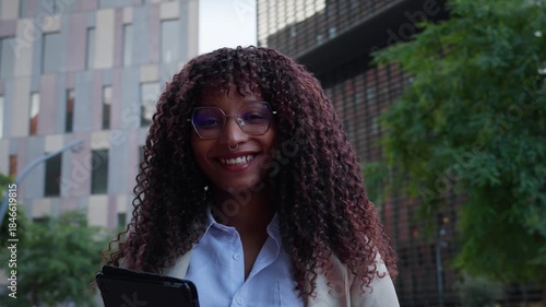Smiling African business woman with glasses holds a tablet and faces the camera in a modern business district. Urban office buildings suggest confidence, technology and leadership.