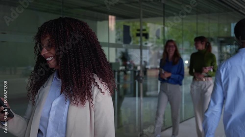 African professional walks through a glass-fronted office lobby while checking her phone, smiling as coworkers pass in the background. Modern workplace vibe with technology, productivity and motion.