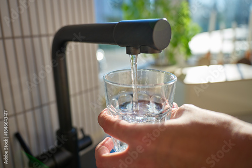 Hand filling glass with drinking water from kitchen faucet in modern home interior. Woman pouring clear water from tap filter. Concept of drinking water, daily hydration and quenching thirst