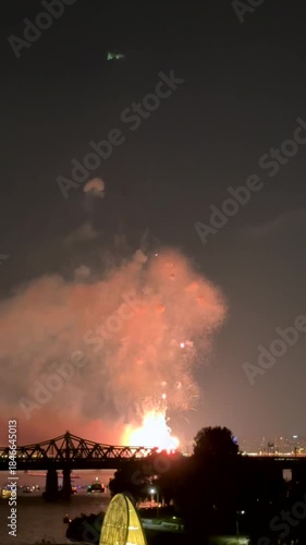 Bright golden fireworks burst above river, lighting up night city and bridge with festive brilliance. Magical celebration vibes shimmer in sky and reflect on water