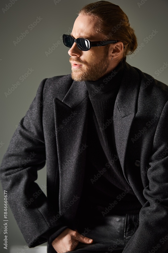 Obraz premium Handsome man in stylish sunglasses and elegant coat poses confidently in grey studio