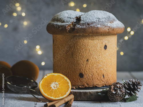 Traditional Italian handmade panettone on a wooden table with orange and cinnamon. Christmas lights with bokeh effect in the background.