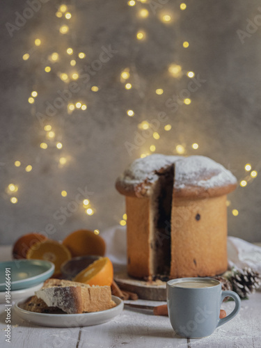 A slice of traditional Italian panettone next to a cup of coffee on a wooden table with Christmas lights. Space for text
