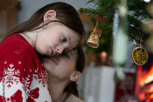 Portrait of mother hugging sad daughter in Christmas sweater near decorated tree, emotional holiday family moment