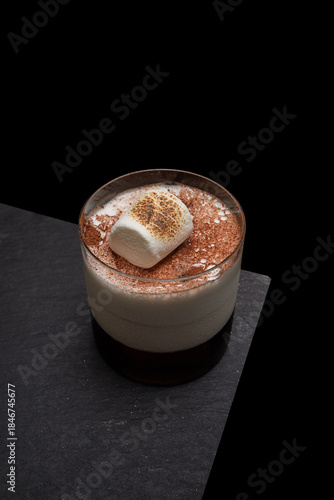 A delicious cocktail garnished with a toasted marshmallow and cocoa powder, artfully presented on a black surface. The drink looks appetizing and elegant.