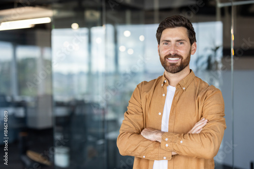 Smiling bearded man standing in a modern office with arms crossed, projecting confidence, professionalism and success as a young executive or entrepreneur in a corporate setting
