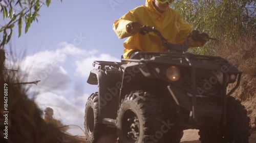 Slow motion. Low angle sand gully quad rider emerges in yellow jacket, camera close to ground as sand sprays, sunlight through willow branches, hands on handlebars, slow