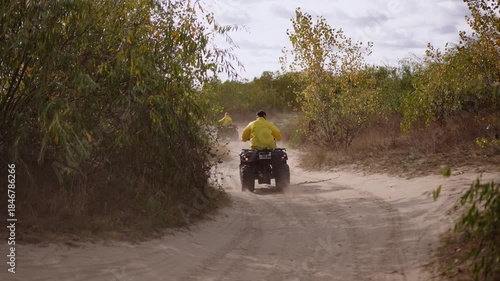 Yellow jacket rider navigates winding dunes, twoquad convoy threading leafy scrub corridors and soft sand tracks under overcast sky, steady pace and scenic