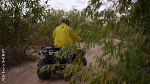 man in yellow jacket riding ATV down sandy trail, dust kicking up behind tires, dense green shrubs lining narrow path, serene overcast sky, engine rumble