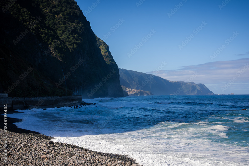 Naklejka premium Steep forested cliffs rise over a pebble beach and Atlantic surf on Madeira's north coast, Portugal. Terraced villages dot slopes, a headland curves, midday light defines
