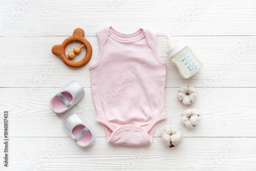 Baby bodysuits and eco friendly accessories - booties, wooden rattle toy and baby bottle with milk on white wooden background, top view