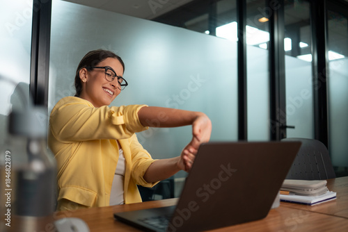 Professional woman in yellow blazer stretching arms during work break at desk in modern corporate office workspace