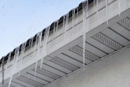 Photo of multiple icicles formed on the roof of house. Big sharp dangerous icicles on the roof right over sidewalk. Winter weather conditions concept. Icicle danger in cities concept.