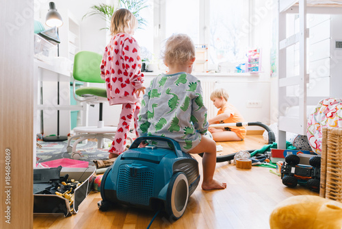 Children's room. Children elementary ages playing and cleaning in messy nursery. Boy vacuums the room while others siblings play. Child's playroom full of toys. Family at home.