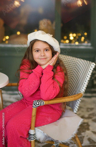 Adorable little girl in a festive winter setting, sitting on a vintage bench and holding a warm cup. Child wearing a bright pink cozy outfit and a white beret, smiling gently at the camera. Christmas