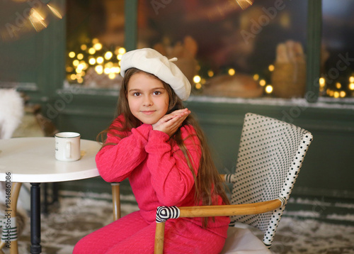 Adorable little girl in a festive winter setting, sitting on a vintage bench and holding a warm cup. Child wearing a bright pink cozy outfit and a white beret, smiling gently at the camera. Christmas
