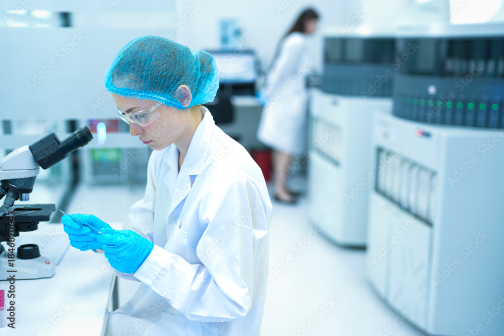 Fototapeta premium Young Caucasian female scientist in safety gear using micropipette and microscope in modern sterile lab. Focused research with colleague in background for medical science.