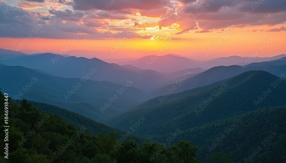 Fototapeta premium Hills and valleys fade into misty blue haze under vibrant sunset sky. Mountain layers glow with orange pink hues, tree covered slopes stand dark green.