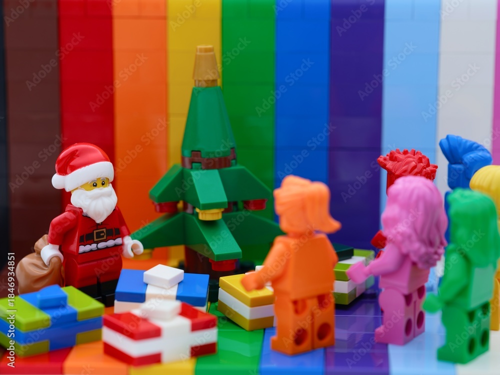 Naklejka premium Tambov, Russian Federation - October 19, 2025 Lego Santa Claus standing near Christmas tree with gifts in front of rainbow monochrome minifigures. Rainbow background