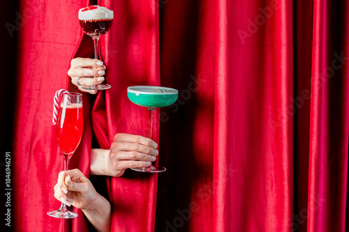 Three festive, colorful holiday cocktails, including one with a candy cane, are presented by hands appearing through luxurious red velvet curtains