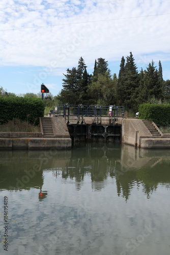 The photo shows a lock in a river set in a Mediterranean landscape, surrounded by lush green vegetation in summer and bathed in warm sunlight.