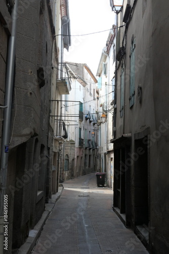 The photo shows a narrow street in a Mediterranean medieval town, with old stone buildings, warm earthy colors, and sunlight filtering between the close-set walls.