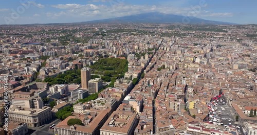 Aerial view of the city center of Catania, Sicily, southern Italy. In the background, on the horizon, Mount Etna looms in silhouette, dominating the panorama. It is a sunny summer day.
