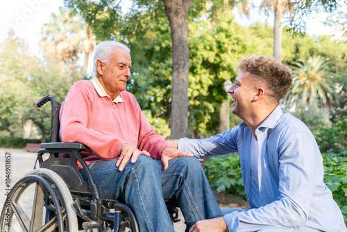 Obraz na płótnie Smiling senior man in wheelchair with young adult grandson at park