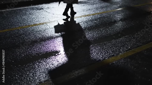 A Woman Walking on a Wet City Street at Night Under Streetlights With Yellow Lines Reflecting in Puddles Dramatic Shadows Cast by the Sole Figure.