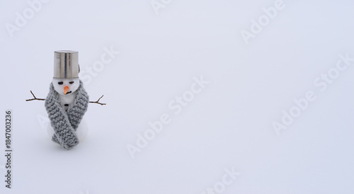 Small snowman with bucket hat and knitted scarf standing in white snow