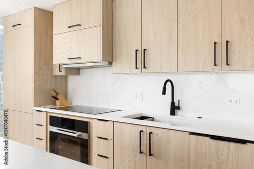 A kitchen detail with a black faucet on a white marble countertop, stovetop and stainless steel oven, and modern wood cabinets.
