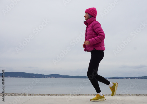 Outdoor light running workout on beach in cold weather, woman jogging in jacket and hat, full body view, simple daily exercise, healthy habits, active lifestyle, winter wellness routine.