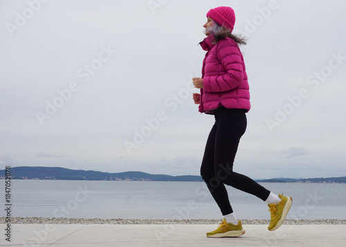 Outdoor light running workout on beach in cold weather, woman jogging in jacket and hat, full body view, simple daily exercise, healthy habits, active lifestyle, winter wellness routine.
