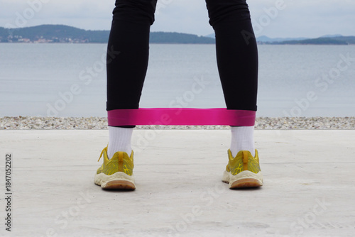 Outdoor resistance band leg exercise on beach in cold weather, legs in sneakers view from behind, simple daily rehabilitation workout, recovery training, balance, mobility, wellness routine.