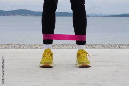 Outdoor resistance band leg exercise on beach in cold weather, legs in sneakers front view, simple daily workout, everyday training, balance, mobility, wellness routine. Healthy habits.