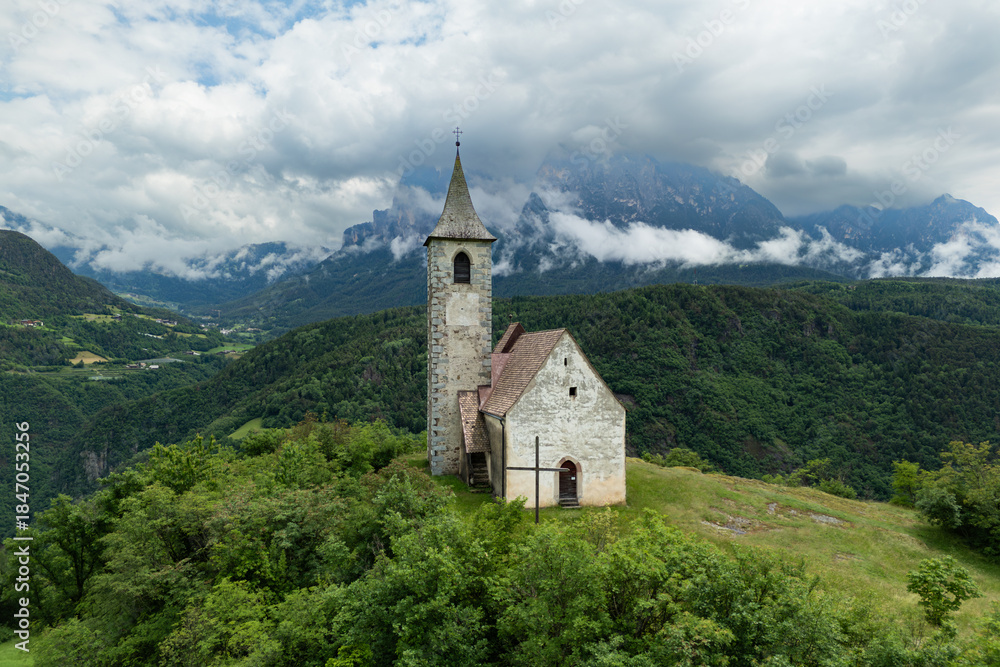 Fototapeta premium Chiesa di Sant Andrea in Renon above a hillside pasture with tree clusters, Catinaccio backdrop, aerial view. Dolomites region