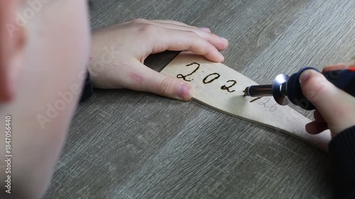 A childs hand writes by burning wood, pyrographic New Years text 2025/26, over shoulder shot