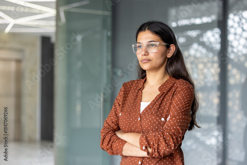 Free Download Young adult indian businesswoman wearing glasses and a polka dot shirt, standing with arms crossed, thoughtfully looking away in a modern office, representing confidence and success - GFXLoad
