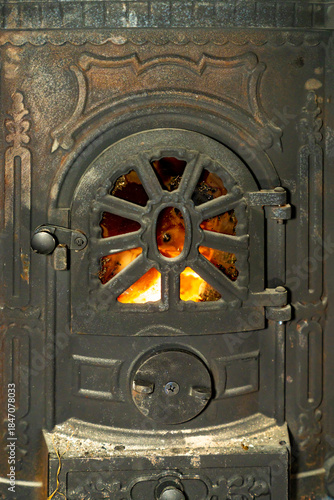 Cast iron wood burning stove door with visible fire flame behind metal grate. Indoor heating appliance close up photography. Home heating and solid fuel stove concept.
