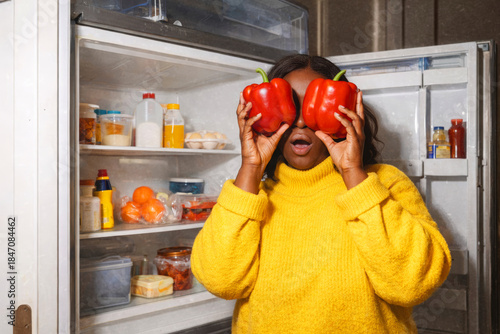 Playful Black woman in a yellow sweater, holding red bell peppers in front of her eyes while standing by an open refrigerator. Healthy eating, food choice, vegetarian lifestyle, nutrition awareness.